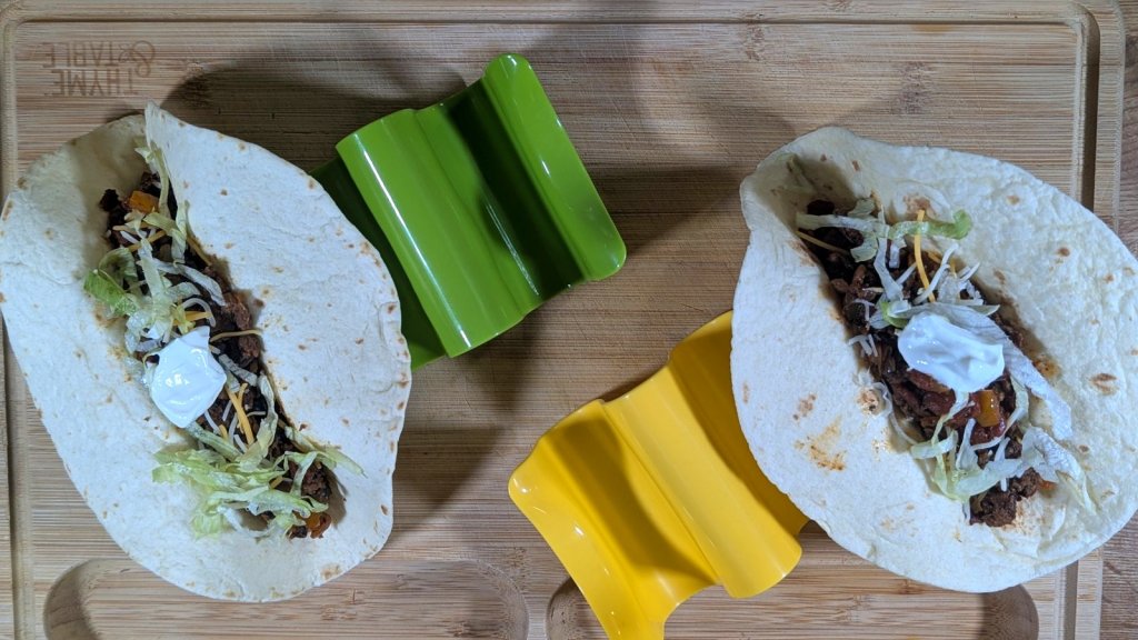 Two soft tacos on a bamboo cutting board, one with ground beef and one with black beans and lentils, topped with lettuce, cheese and sour cream, next to green and yellow taco holders